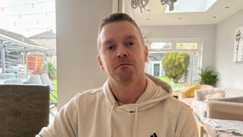 A man with short dark hair and a stubbly beard wears a beige hoodie and sits in a house setting with chairs and sofas behind him and a large window to his garden over his right shoulder. 
