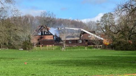 Children's nursery, Tigers in a L-shaped barn conversion. The building is dark brown wood with small windows on first floor and velux windows in its roof. A fire engine is to the right with water being sprayed onto the building.