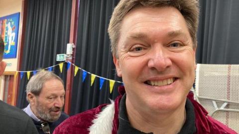 Jeremy Fish smiles at the camera inside a community hall. He has short dark hair and is wearing a red and white Santa Claus top. Behind him other volunteers at a wrapathon event can be seen talking.