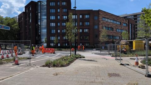 A foot way leads towards a multi-storey office building. The road next to the path is blocked off for road works, with people in orange high-vis working on it.