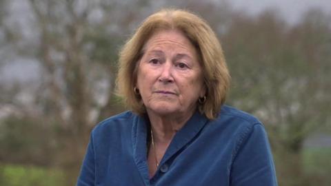 A woman with ginger hair wearing a blue jacket with some trees in the background