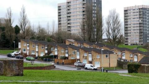 Tower blocks are seen rising up behind a row of houses at Druids heath. There are open spaces with grass and trees dotted around the landscape. Cars are parked in the road outside the houses.