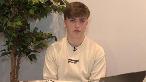 A teenage boy with short brown hair and a long sleeve t-shirts sits at a desk with a laptop