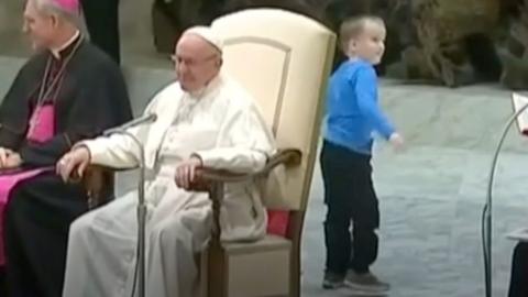 A little boy stood behind the Pope, who is sat in a chair