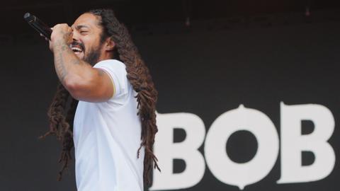 Bob Vylan frontman Bobby Vylan performing at Glastonbury Festival. He is wearing white polo shirt and has several tattoos on his arms. The word 'Bob' is behind him on the stage.