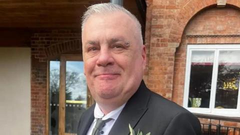 A man with short cropped grey hair and a ring piercing on the end of his left eyebrow. He is wearing a black blazer over a white shirt and a grey tie. There is a flower in the right breast pocket. He is standing in front of a red-bricked building and smiling.
