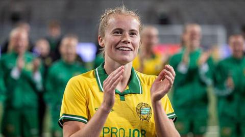 Close-up image of Julie Blakstad during Hammarby IF's Women's Champions League qualifying round match against Metalist 1925 in August 2025. She has fair hair, tied back, and is wearing a yellow shirt which has a green collar and trim on the sleeves. She is clapping.