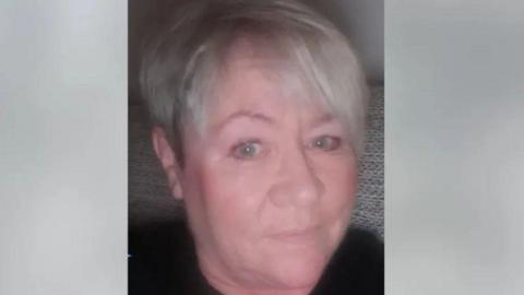 Kelly smiles a small smile in a selfie. She is sat on a sofa and has short grey hair. 