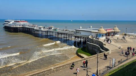 We can see Cromer Pier on a sunny day. There are people walking on the promenade.