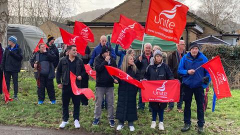 A group of workers waving red Unite UNION flags on the roadside. They are all wearing dark clothes and dressed for the cold weather. One person can be seen using a whistle and another person is holding a brass horn.