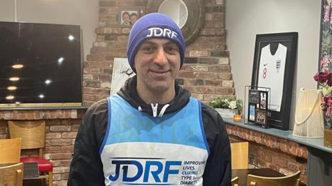 Mid shot image of Mehdi Taheri. Mr Taheri is wearing a black long sleeve jacket with a blue and white vest which states: "JDRF on the front". He is also wearing a purple beanie with a white JDRF logo on the front. 