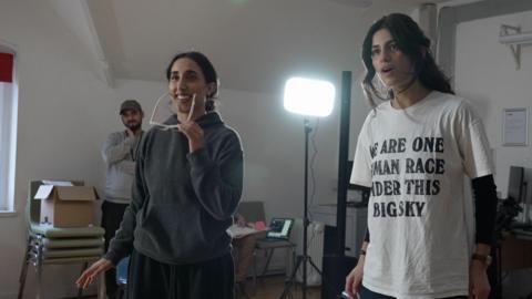 Two female actors in a rehearsal room - one in a grey hoodie , taking off white sunglasses and another wearing a white t-shirt with the words 'We are one human race under this big sky'