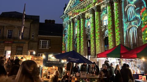 It is evening. An array of market stalls at night time. There are tens of people moving around them. In the background a large pillared building is illuminated with Christmas imaging, including wreaths.