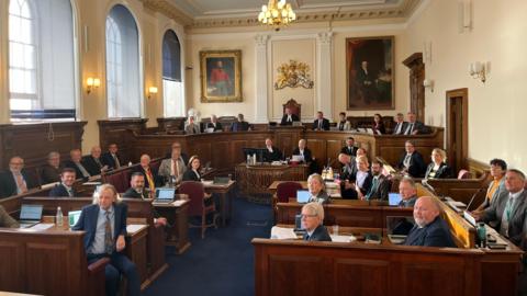 There are 38 politicians sitting at wooden benches all looking beyond the camera in Guernsey's Royal Court