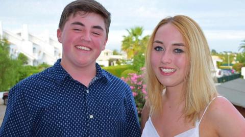 Photo of Scott Allan and his sister Katie together before her conviction. It looks like a holiday shot. Scott is just 14 but he's taller than his older sister. He is smiling at the camera, wearing a blue shirt with small star patterns. He has short dark hair. His sister is also smiling at the camera. She is wearing a white sun dress. She has blonde hair and blue eyes. The background appears to white apartments and trees of a holiday resort.