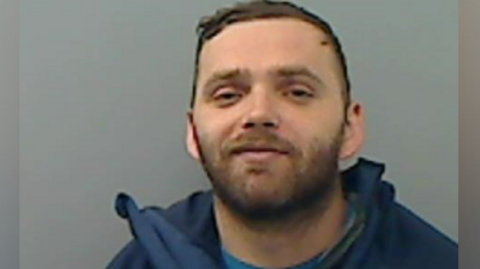 A man with short brown hair, a brown beard, smirks at the camera, in a police mugshot. The background is a painted grey wall and he is wearing a blue t-shirt and navy jacket. 