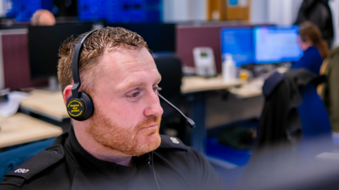 A man  with short ginger hair and bead with a headset on in the police call centrere