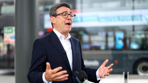 Man standing in front of a mic in a bus station. his hadns are out stretched, he's wearing black glasses, dark blue blazer and white shirt.