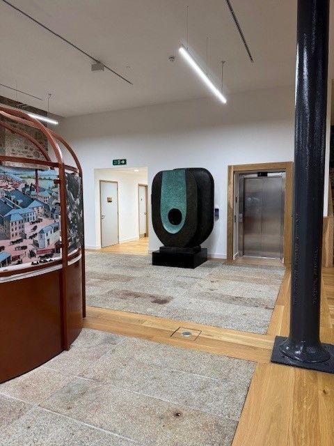 The sculpture sitting the a room with a granite and wooden floor. The sculpture is as large as a doorway and is an oval shape mounted on a plinth. It is black with a green oval shape and a round hole half way down it.