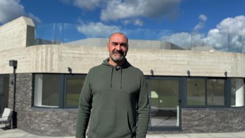 A smiling man wearing a green hoody, standing in front of a black and white building. 