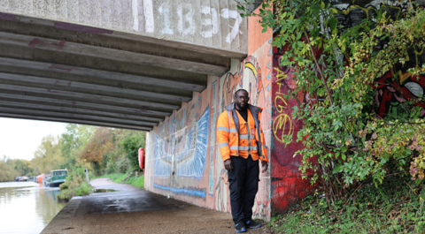 Devon standing by a canal on an overcast day. He is wearing an orange vis jacket. There is greenery growing near him. There is graffiti on the wall behind him. A boat can be seen moored in teh distance.
