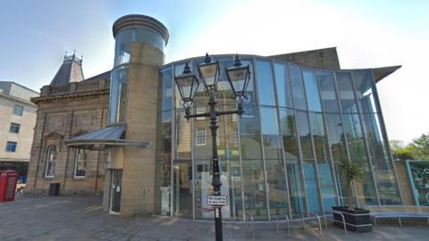 A Google Street View screenshot of the front of Sunderland's Museum and Winter Gardens. The ornate Victorian stone building has a large modern curved glass entrance. A Victorian style black metal streetlight, made up of three lamps resembling gas lamps, sits in front of the building.