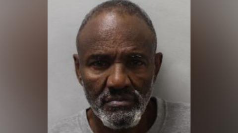 Police custody photograph of Musse Ali, a man with short grey hair, a short grey beard, and a grey crew-neck top.