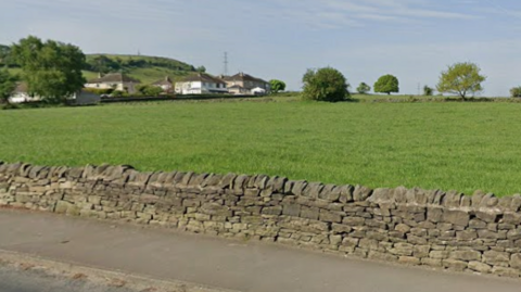 A large green field contained by a dry-stone wall. In the background, residential houses can be seen.