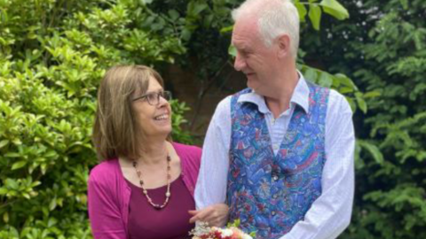 A woman with bobbed brown hair and glasses, wearing a pink top and cardigan and pink necklace. She is looking at, and has her arm lopped through that of, a man with white hair, wearing a pale blue shirt and patterned blue waistcoat. he is also looking at her. They are stood in front of a large green bush.