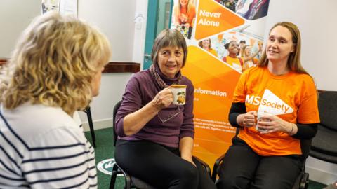 Three women sit together over a cup of tea. The woman on the right is wearing an orange tshirt that says 'MS Society'. To her left is a woman wearing a purple jumper. They are both looking at a third woman to the left of frame in a black and white striped top. They appear to be talking. In the background there is a standing poster for the MS Society.