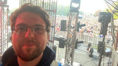 An early thirties male with a beard and glasses, standing back stage at a festival in front of a large crowd.