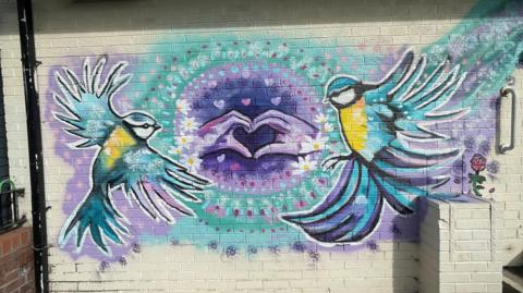 A mural showing two birds with yellow chests, blue wings and white faces against a blue and purple background. The image also features two hands making a heart shape with their fingers in the centre of the artwork. 