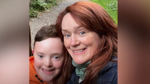 Finn has brown hair and wearing an orange hoodie. Angela has red hair and wearing a navy jacket with a green scarf. They are standing on a path surrounded by trees and smiling at the camera.