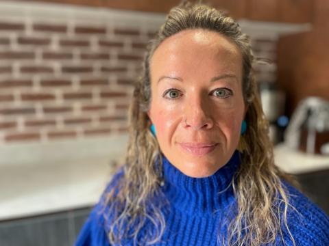 Laura is a white woman with long, fair wavy hair. She is wearing a blue jumper and is stood in the kitchen of her home.