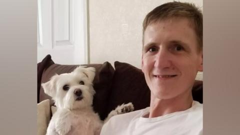 A man with light brown hair and wearing a white T-shirt sits on a brown sofa with a white terrier dog sitting next to him.