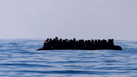 A inflatable dinghy carrying around 65 migrants can be seen silhouetted against a grey sky on a blue sea, as it crosses the English Channel in March