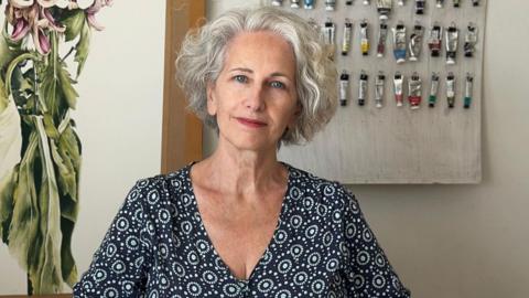 Marie Burke is sitting in front of a wall with paints hanging from nails. She is wearing a patterned shirt and has grey hair.