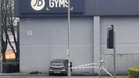 A silver or grey car has crashed into the wall of a JD Gym. There is a hole in the wall.