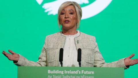 Michelle O'Neill standing at a green lecturn with a green background behind her. She is motioning with her hands. She is wearing a cream jacket and white shirt. She has blonde hair, tied back.
