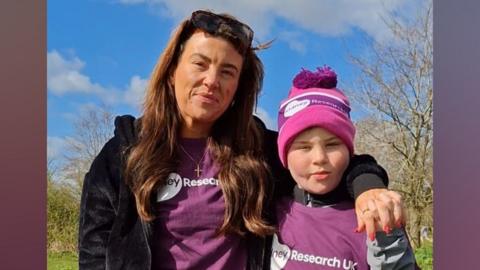 Sammy, a woman with shoulder length dark brown hair, with Caelan, a boy wearing a purple T-shirt and woolly hat. They are on their walk at Ferry Meadows in Peterborough.