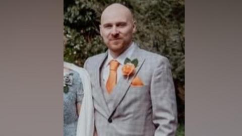 Gareth Underwood, who is bald with a light brown goatee and it wearing a light grey suit with a peach tie and a matching flower in his lapel. 