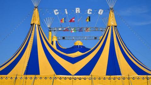 A blue and yellow circus tent with multiple country flags and the word circo in capitals above it, set against a bright blue sky.