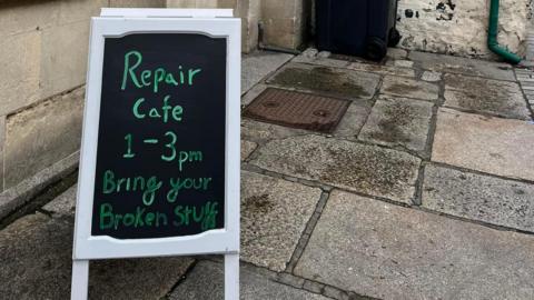 A chalk A-board sign outside a stone building that says 'Repair cafe 1-3pm, bring your broken stuff'