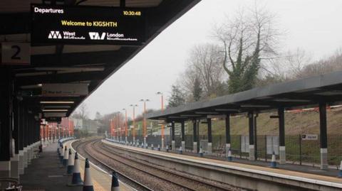 A platform at a railway station with two tracks. The platform is coned off. A departure board says welcomd to KIGSHTH.