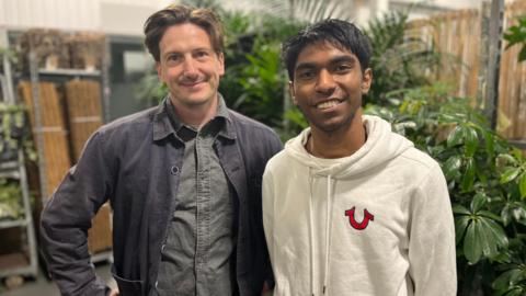 Two men smile at the camera in front of plants. One is in dark clothing, another in a white hoodie