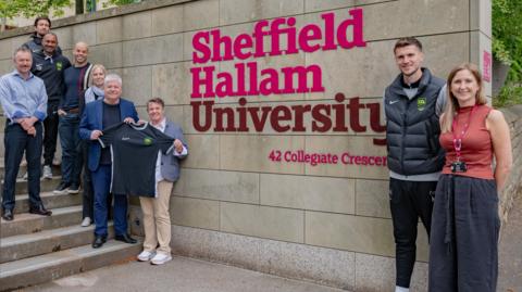 Six men and three women are standing either side of a large Sheffield Hallam University sign which is red and pink lettering on a stone wall.  Seven are standing to the left-hand side, up a set of steps. Two are holding a black football shirt with a grey neckline