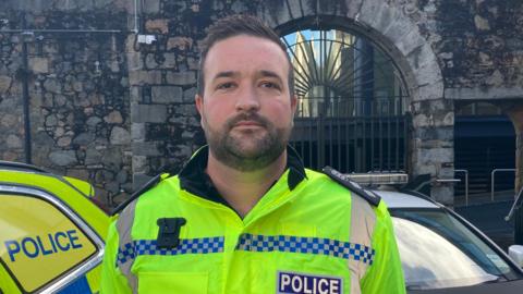 Insp Asa Flatres in his police uniform, stood in front of two police cars. He has short brown hair and a beard. 
