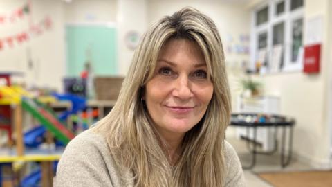 A head and shoulders shot of a woman with long blonde hair, wearing a grey jumper, sitting in a classroom