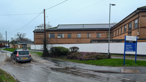 A grey car drives through floodwater on a road outside a hospital. There is a blue sign on the right hand side of the image, indicating it is an NHS site. There is a flower bed on a grass verge at the side of the road and brick buildings in the background.