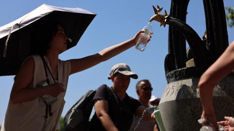 People wearing caps and holding umbrellas fill water bottles at a fountain in London on Monday against a bright blue sky.